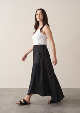 Woman wearing a black satin tiered midi skirt with a white tank top and black sandals against a beige background