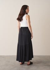 Woman wearing a black satin tiered midi skirt with a white tank top and black sandals against a beige background