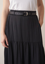 Close up of woman wearing a black satin tiered midi skirt with black belt and a white tank top against a beige background