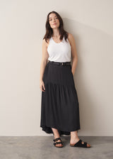 Woman wearing a black satin tiered midi skirt with a white tank top and black sandals against a beige background