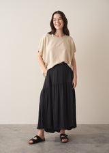 Woman wearing a black satin tiered midi skirt with a cream short sleeved sweater and black sandals against a beige background