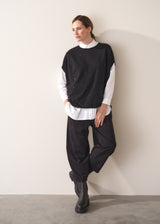 Person wearing a black sweater vest with sequin detail, white shirt, and black trousers, leaning against a plain wall.