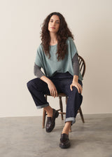 Woman wearing a grey metallic long-sleeve top and dark jeans against a beige background