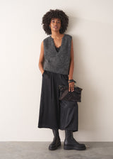 Woman wearing a grey sleeveless sparkle knit sweater and black wide leg satin trousers against a plain background