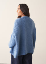 Woman wearing chunky bright blue knit cardigan with patterned blouse and denim trousers against a plain background.