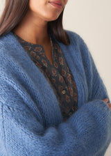 Woman wearing chunky bright blue knit cardigan with patterned blouse and denim trousers against a plain background.