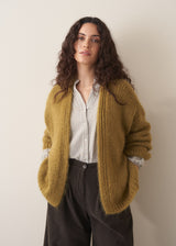 Woman wearing an olive green chunky knitted cardigan over a waffle shirt, against a plain background