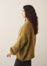 Woman wearing an olive green chunky knitted cardigan and brown trousers, against a plain background