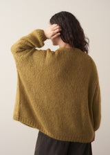 Woman wearing an olive green chunky knitted cardigan and brown trousers against a plain background