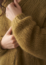 Close up of woman wearing an olive green chunky knitted cardigan against a plain background