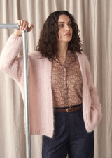 Woman wearing a blush pink chunky knit cardigan with blue denim trousers and floral blouse, against a fabric backdrop
