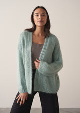 A woman model wears a mint green chunky knit cardigan with a striped top and black wide-leg trousers, against a beige background