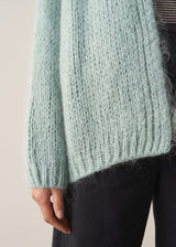 Close up of a woman wearing a mint green chunky knit cardigan with a striped top and black wide-leg trousers against a beige background