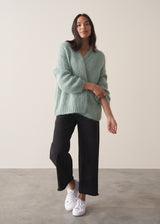 A woman wears a mint green chunky knit cardigan with black wide leg trousers and white trainers, against a beige background