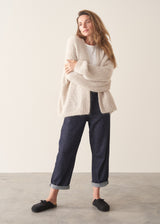 A woman wearing a chunky knit open-front cardigan in a warm cream or oatmeal color, layered over a white top and dark blue jeans, posing against a plain light beige studio background.