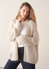 A woman wearing a chunky knit open-front cardigan in a warm cream or oatmeal color, layered over a white top and dark blue jeans, posing against a plain light beige studio background.