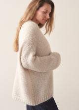 A woman wearing a chunky knit open-front cardigan in a warm cream or oatmeal color, layered over a white top and dark blue jeans, posing against a plain light beige studio background.