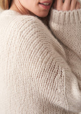 A woman wearing a chunky knit open-front cardigan in a warm cream or oatmeal color, layered over a white top and dark blue jeans, posing against a plain light beige studio background.