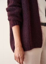 A woman wearing a chunky, fuzzy dark plum purple cardigan over a white V-neck top and white trousers, posing with her arms crossed against a plain off-white wall.