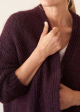 A woman wearing a chunky, fuzzy dark plum purple cardigan over a white V-neck top and white trousers, posing with her arms crossed against a plain off-white wall.