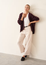A woman wearing a chunky, fuzzy dark plum purple cardigan over a white V-neck top and white trousers, posing with her arms crossed against a plain off-white wall.