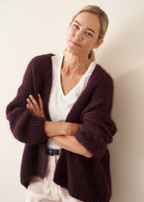 A woman wearing a chunky, fuzzy dark plum purple cardigan over a white V-neck top and white trousers, posing with her arms crossed against a plain off-white wall.