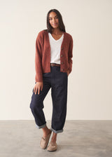 A woman wearing a rust-colored knit cardigan over a white V-neck top and dark blue jeans, holding a beige tote bag against a plain off-white wall.