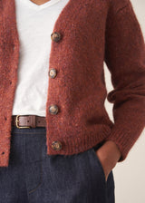 A woman wearing a rust-colored knit cardigan over a white V-neck top and dark blue jeans, holding a beige tote bag against a plain off-white wall.