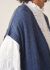 Woman wearing a blue knitted waistcoat, white embroidered top and denim jeans.