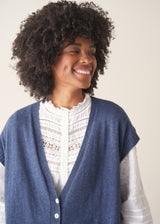 Woman wearing a blue knitted waistcoat, white embroidered top and denim jeans.