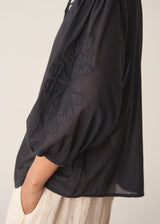 A woman wearing a sheer black embroidered blouse with light linen trousers.