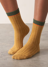 Person wearing heavy knit socks in a mustard yellow wool blend, with retro orange and green ankle stripes, standing on a grey surface