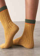 Person wearing heavy knit socks in a mustard yellow wool blend, with retro orange and green ankle stripes, standing on a grey surface