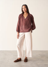 A woman wearing a dark red, brown semi-sheer blouse with a tie necklace and off white trousers.