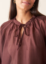 A woman wearing a dark red, brown semi-sheer blouse with a tie necklace and off white trousers.