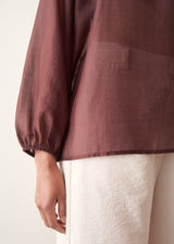 A woman wearing a dark red, brown semi-sheer blouse with a tie necklace and off white trousers.