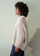 Side profile of a woman in an off white boucle sweater and dark blue jeans against a plain background