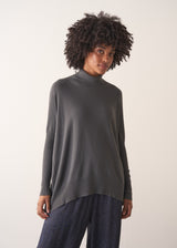 Woman wearing a dark grey turtleneck sweater and blue satin trousers against a plain background