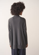 Woman wearing a dark grey turtleneck sweater and blue satin trousers against a plain background