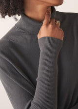 Woman wearing a dark grey turtleneck sweater and blue satin trousers against a plain background