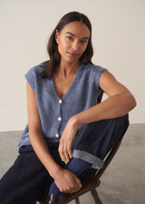 Woman wearing a blue sleeveless knitted waistcoat with blue trousers, sitting on a chair against a plain background