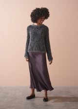 Woman wearing a dark grey fluffy textured sweater with sequin detail, standing against a beige wall