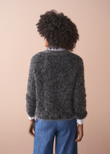 Back of woman wearing a dark grey fluffy textured sweater with sequin detail, blue jeans and collared shirt, facing a beige wall.