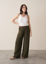 Woman wearing dark green linen wide leg trousers and green waistcoat with neutral sandals against a beige background