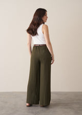 Woman wearing dark green linen wide-leg trousers and a white tank top with neutral sandals, against a beige background