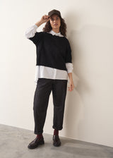 Woman wearing an oversized white shirt with black jumper and dark jeans, paired with brown loafers and cap