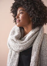 Woman wearing a large textured chunky knit scarf in neutral tone, against a plain background
