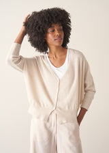 A woman wearing a light oatmeal cream knit cardigan over a white V-neck top and matching light oatmeal trousers.