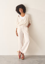 A woman wearing a light oatmeal cream knit cardigan over a white V-neck top and matching light oatmeal trousers.