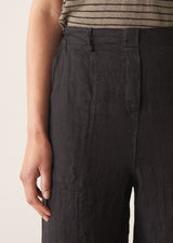 Woman wearing a striped t-shirt and black linen trousers, standing against a plain wall.